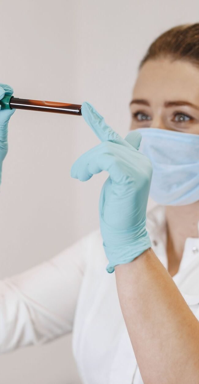 A female healthcare worker wearing gloves and a mask examines a blood sample in a test tube.