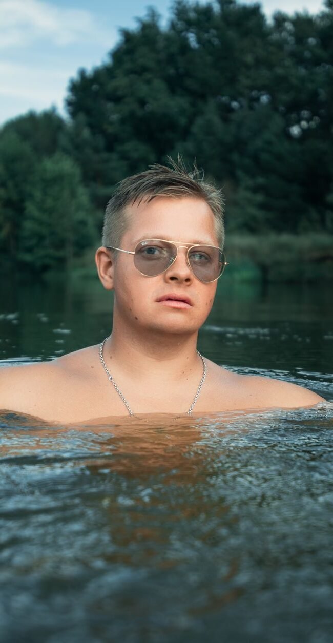 Man with sunglasses enjoys a refreshing swim in a river surrounded by lush greenery.