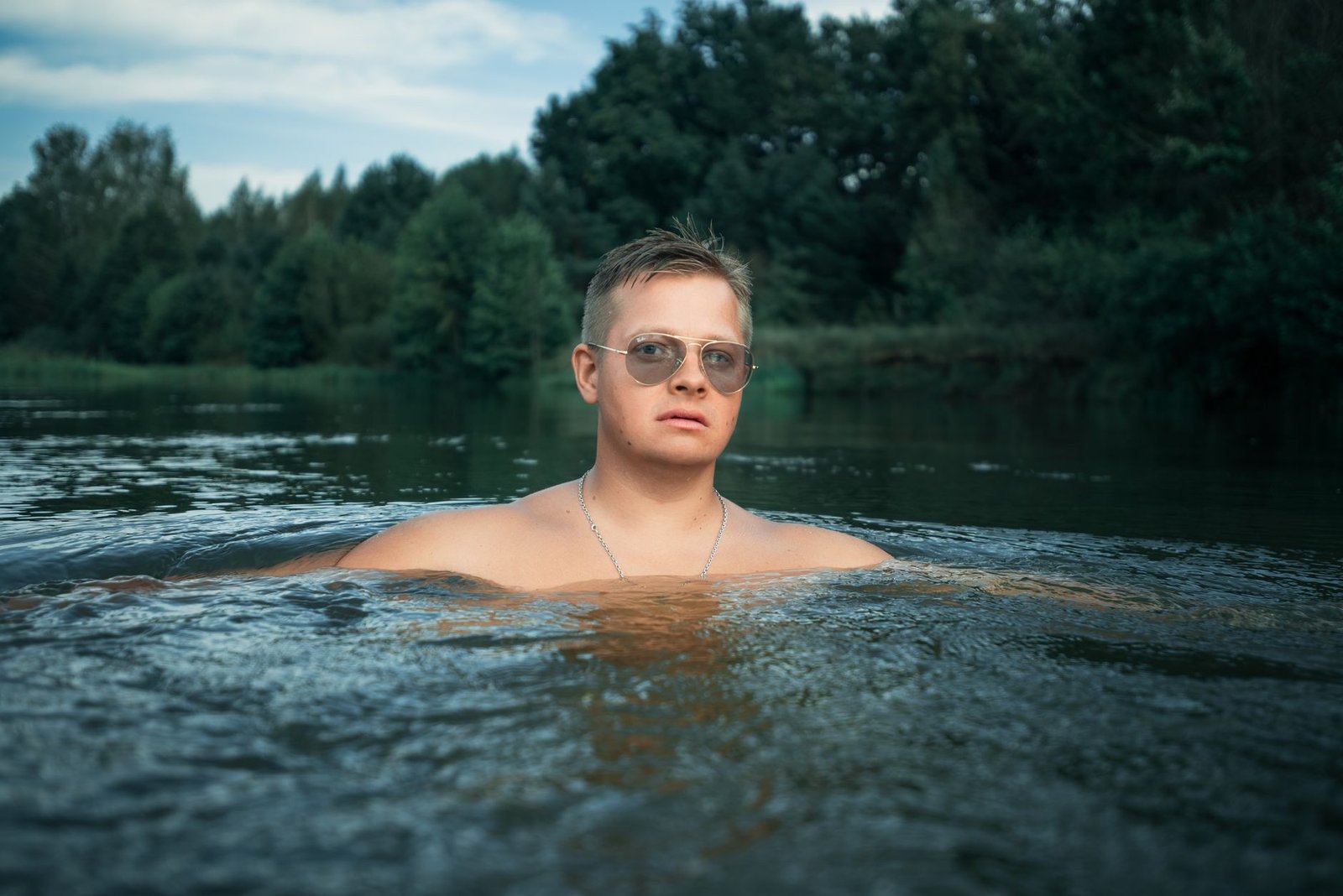 Man with sunglasses enjoys a refreshing swim in a river surrounded by lush greenery.