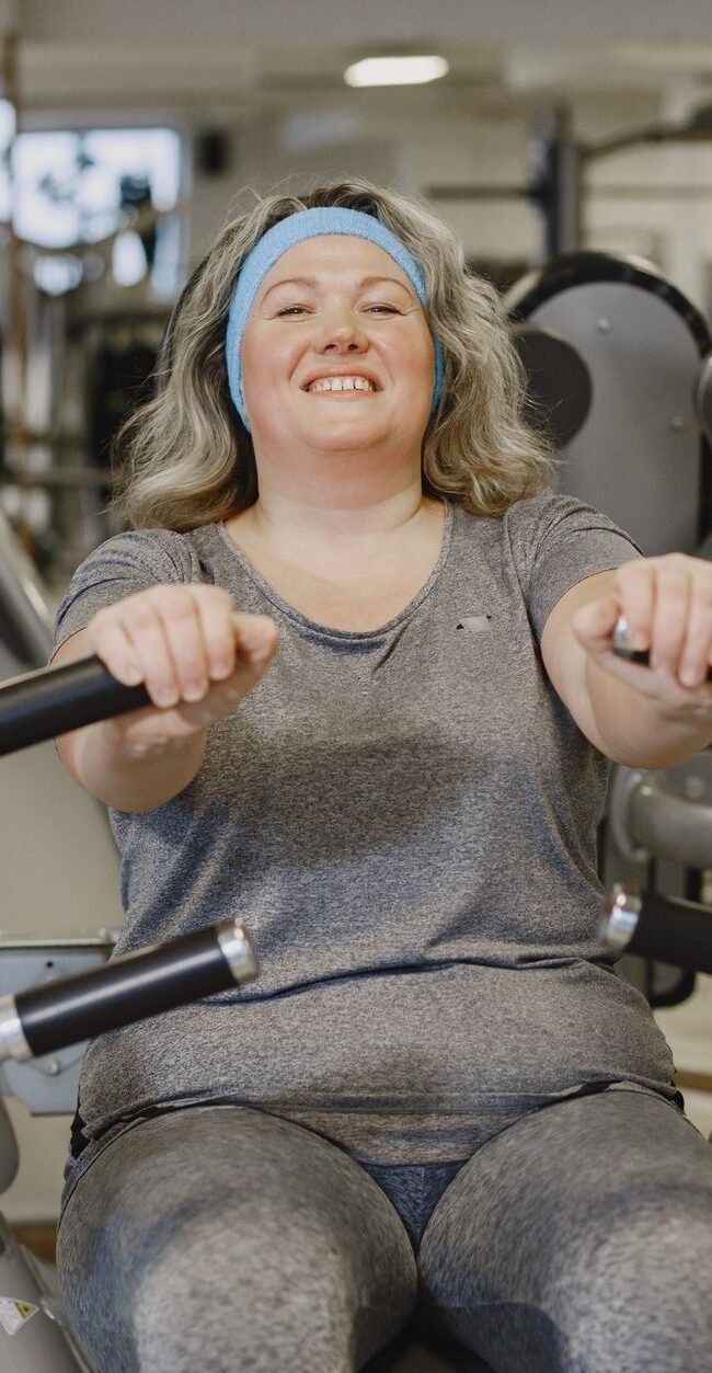 Smiling woman exercises on a gym machine, promoting fitness and well-being.