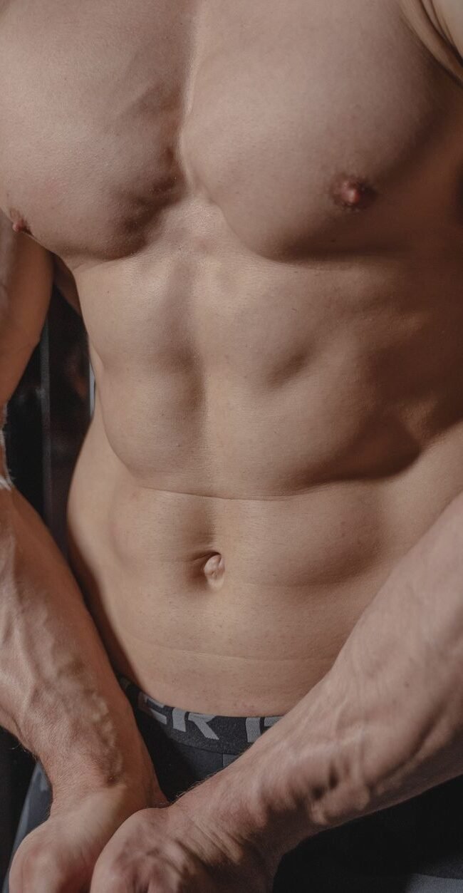 Shirtless male bodybuilder showing muscular torso and defined arms in a gym setting.