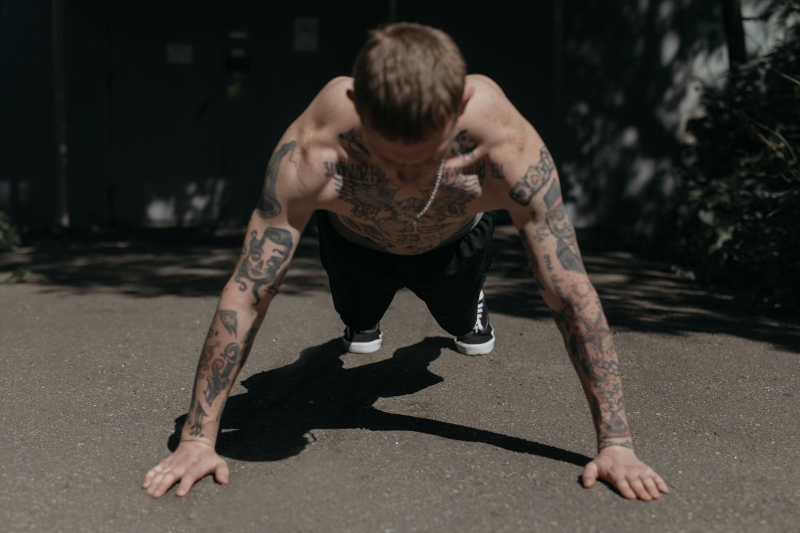 Shirtless tattooed man performing push-ups on a concrete path outdoors, showcasing fitness and endurance.